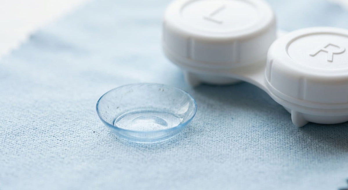 A reusable soft contact lens beside a lens case, with subtle microscopic particles or bacteria around the lens